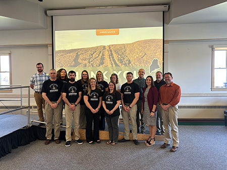 Photo of the Leadership Aroostook Beta Class Graduates. They are lined up in front of a projector with an image of a potato field and text that says, "Thank You." They are wearing Black Leadership Aroostok T-Shirts.