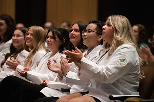 Husson University pharmacy students seated in The Gracie Theatre in Bangor and wearing white coats clap.