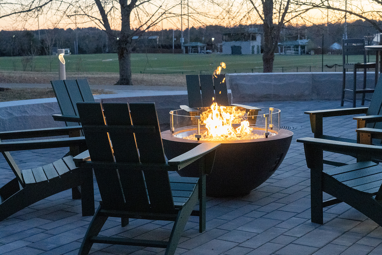A fire burns in a round fire pit with chairs surrounding it and Husson University's campus in the background.