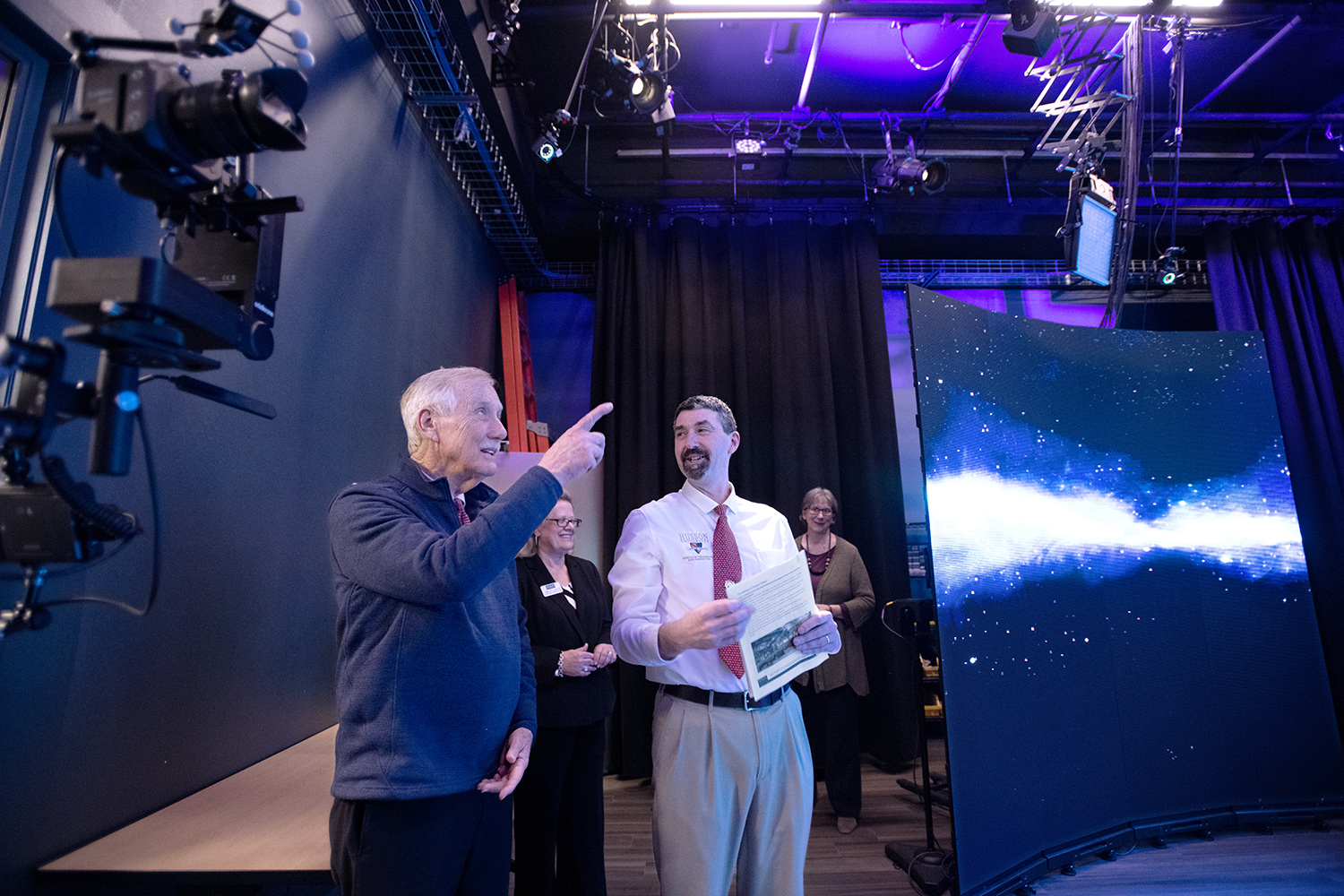Senator Angus King is shown in a technology lab with screens where he is being spoken to by another man.