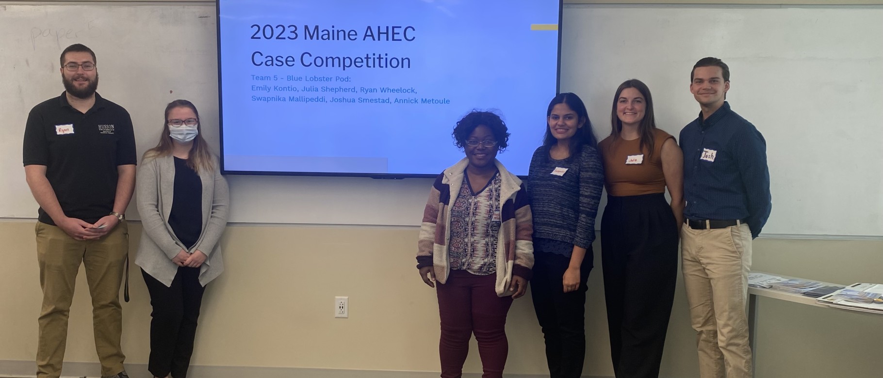 Two people are shown to the left of a screen and four people to the right. On the screen, the words “2023 Maine AHEC Case Competition” are displayed.