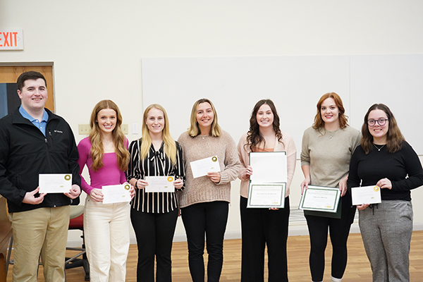 Seven smiling people are lined up in a white room. Each is holding a certificate.