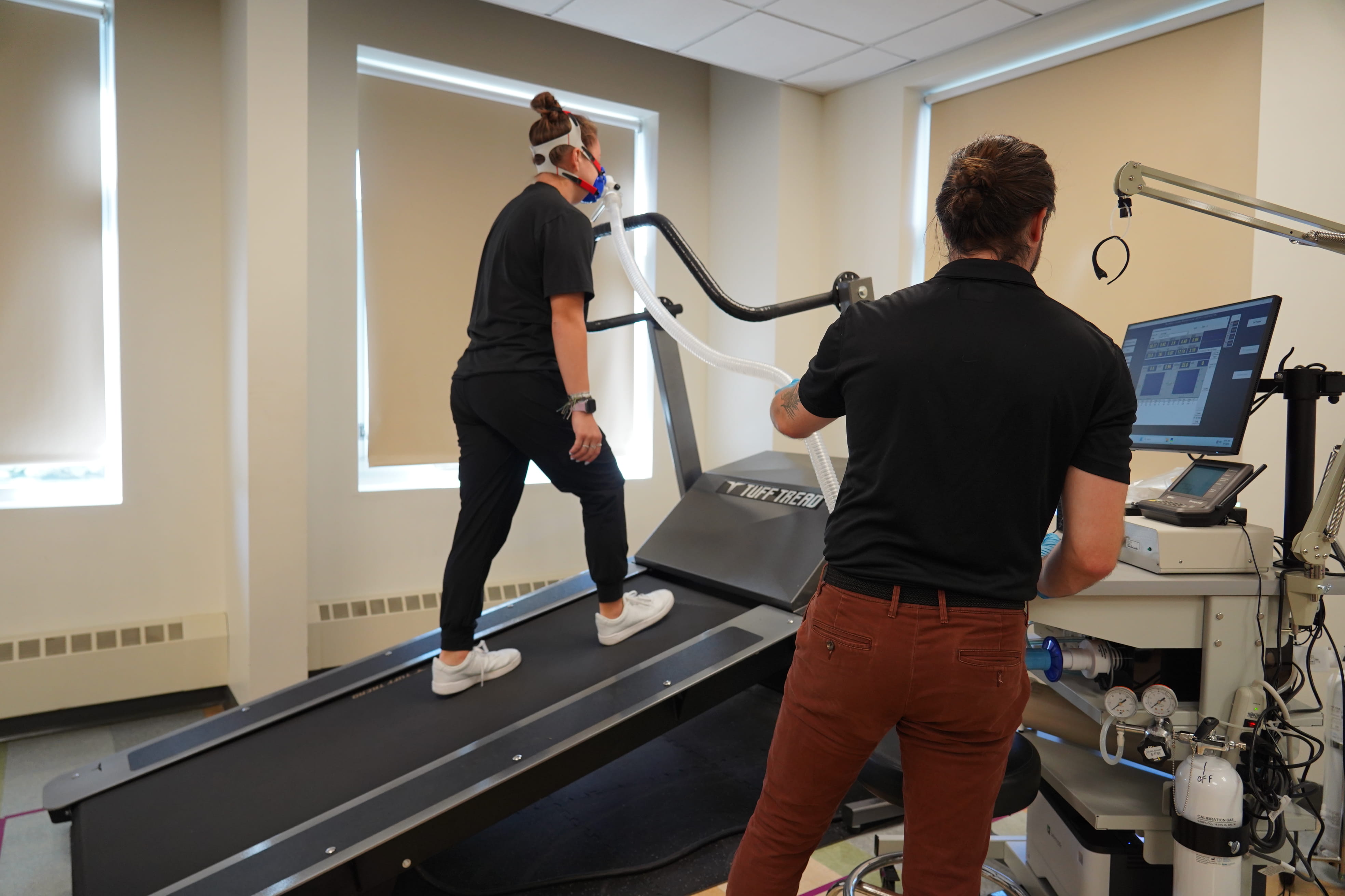An man collects data on a computer while monitoring a woman walking on a treadmill.