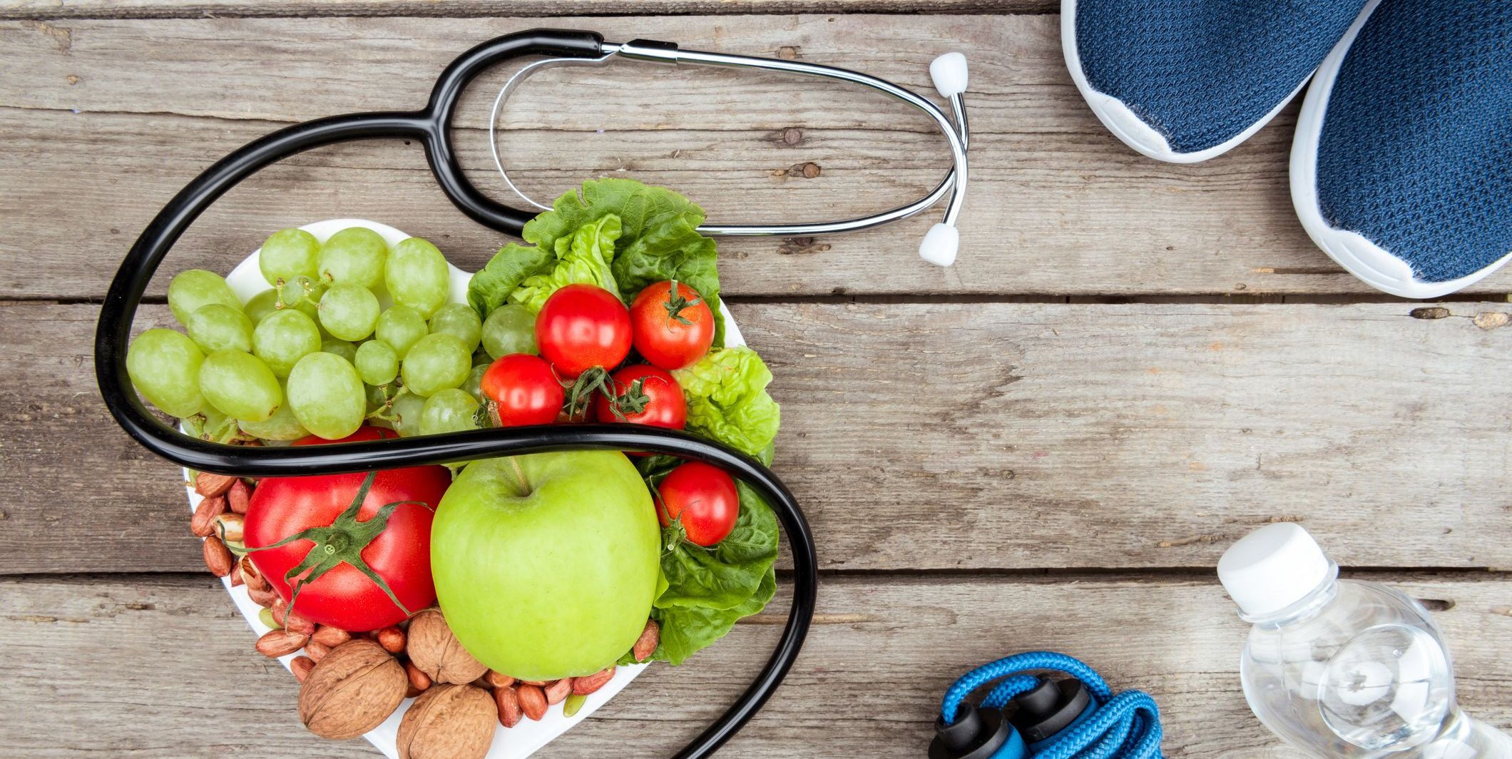 A heart-shaped plate of fruits and vegetables, stethescope, shoes and other workout materials on a wooden table.