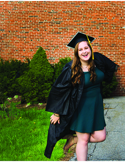 Victoria Green, a Husson communications graduate, poses in front of a brick wall while wearing a graduation gown