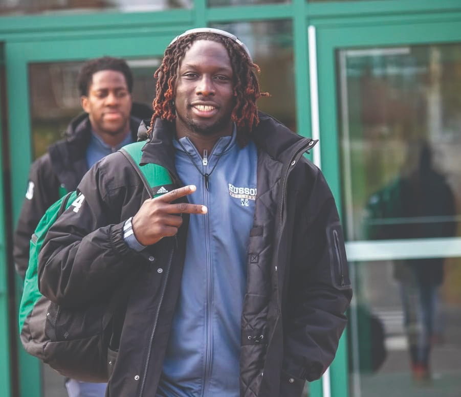 Hamza Abdulrahman, a Husson grad, smiles and gives a peace sign with his fingers while carrying a backpack on one shoulder. 