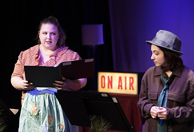 two actresses rehearse play scripts while on stage in front of an "on air" sign