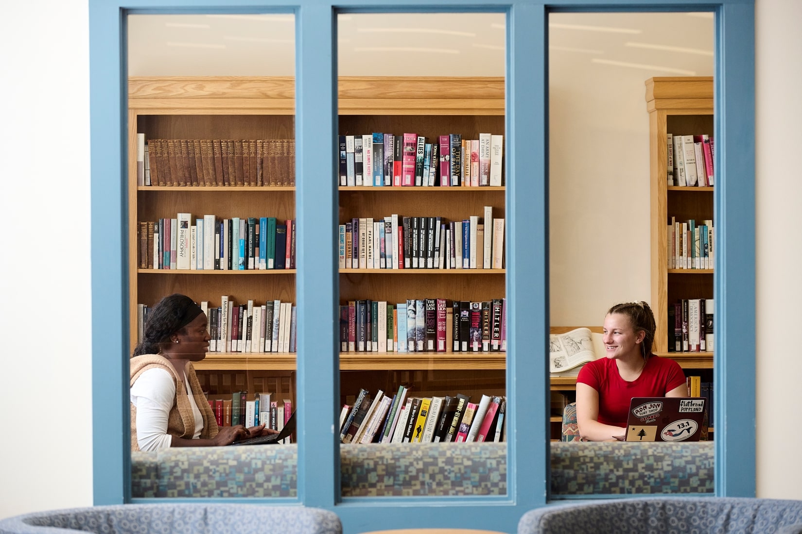 View through a window of a two women talking to each other while sitting in a library.