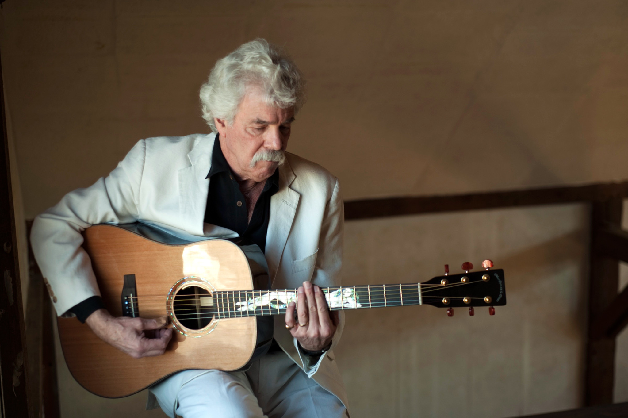 Tom Rush playing an acoustic guitar while sitting on a stool.