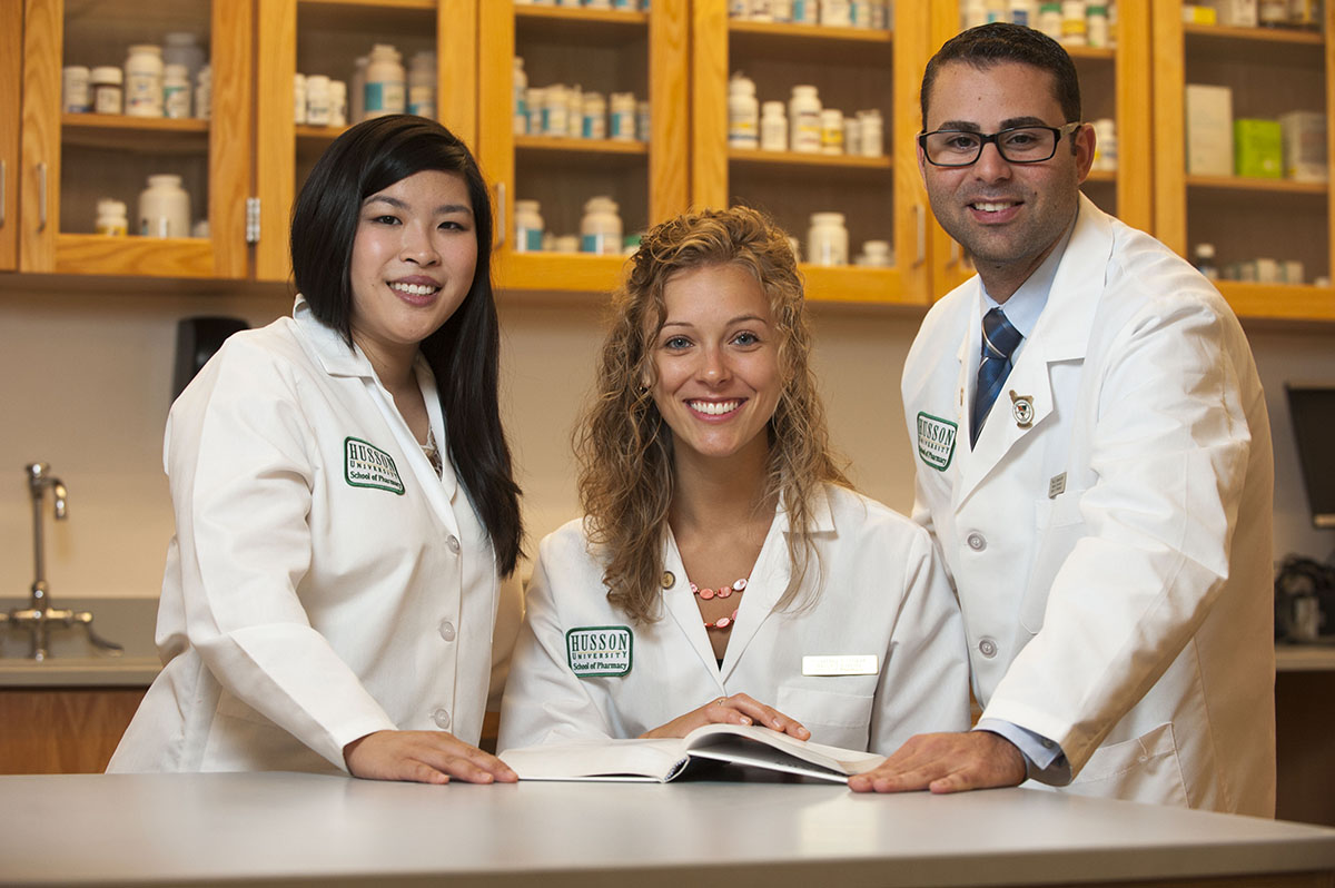 Three smiling pharmacy students standing next to each other behind a table that has a book on it.