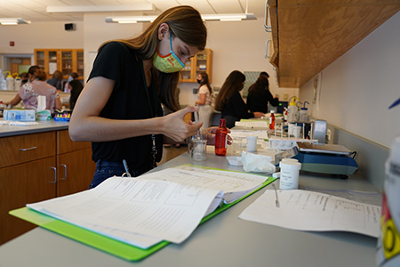 A pharmacy camp student stands over a lab station and injects a syringe of liquid into a beaker.