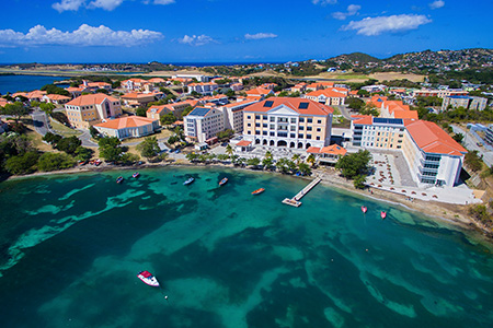 Image is of St. George's University Campus. The campus buildings are tan with orange roofs. The campus is located right on the shore of Grenada in the West Indies. The water is a beautiful aqua blue and there are 8 boats near the shore. 
