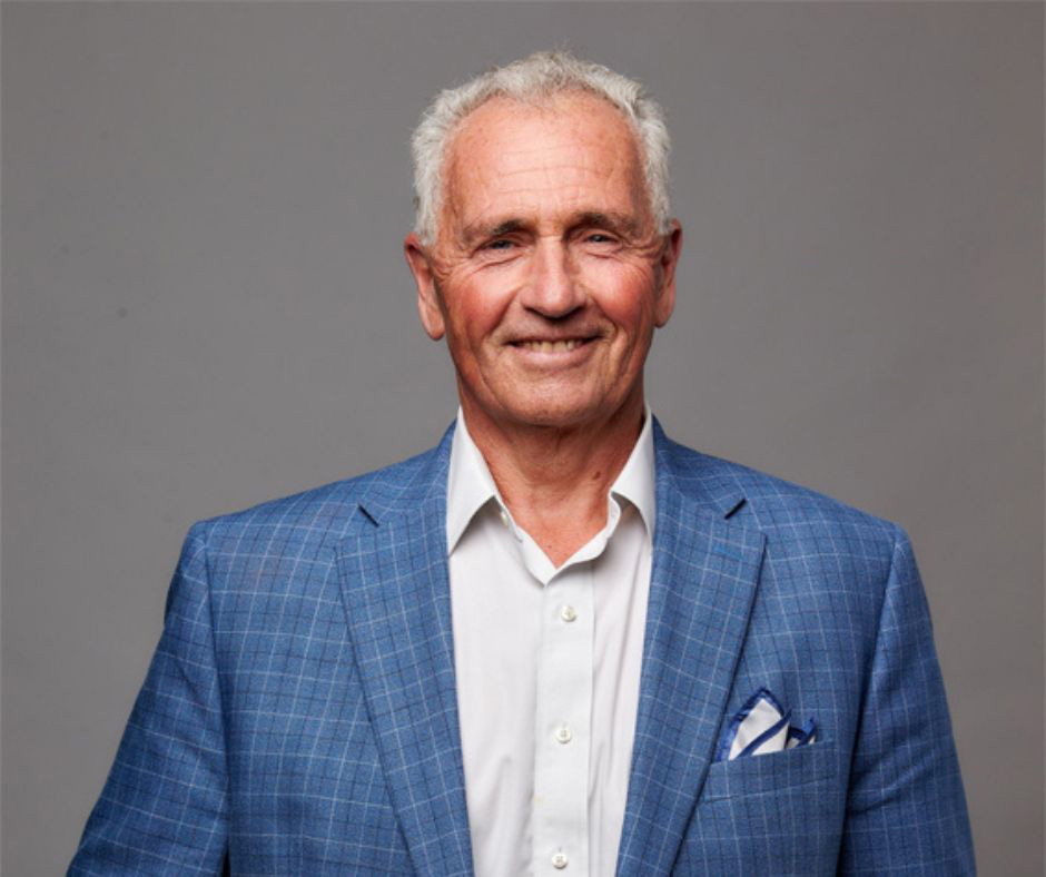 Sean Riley, president and CEO of Maine Course Hospitality Group, poses for a headshot. Riley wears a blue checkered suit with a white shirt and blue and white handkerchief. He poses in front of a grey backdrop.