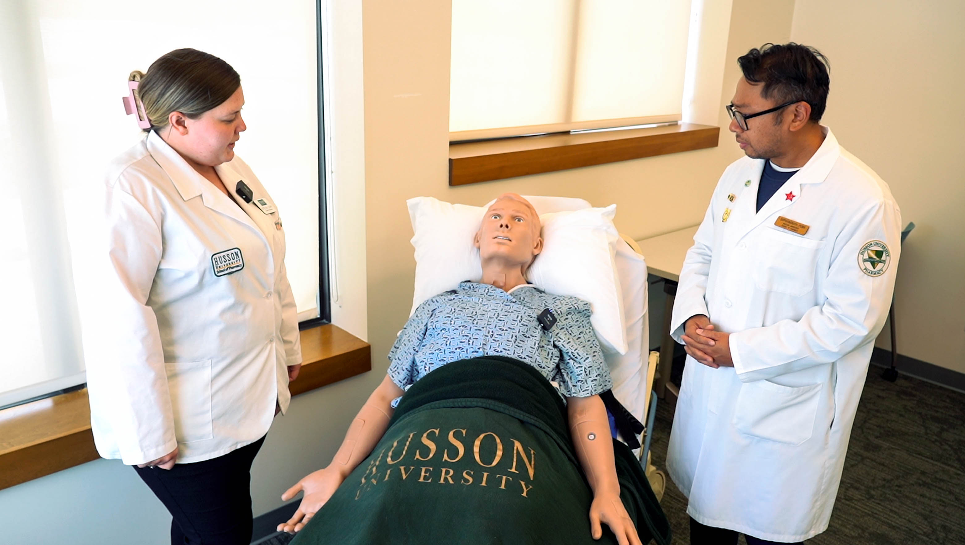 The HAL S5301 interdisciplinary patient simulator manikin is shown wearing a hospital gown on a hospital bed. Two students stand beside the bed on other time.