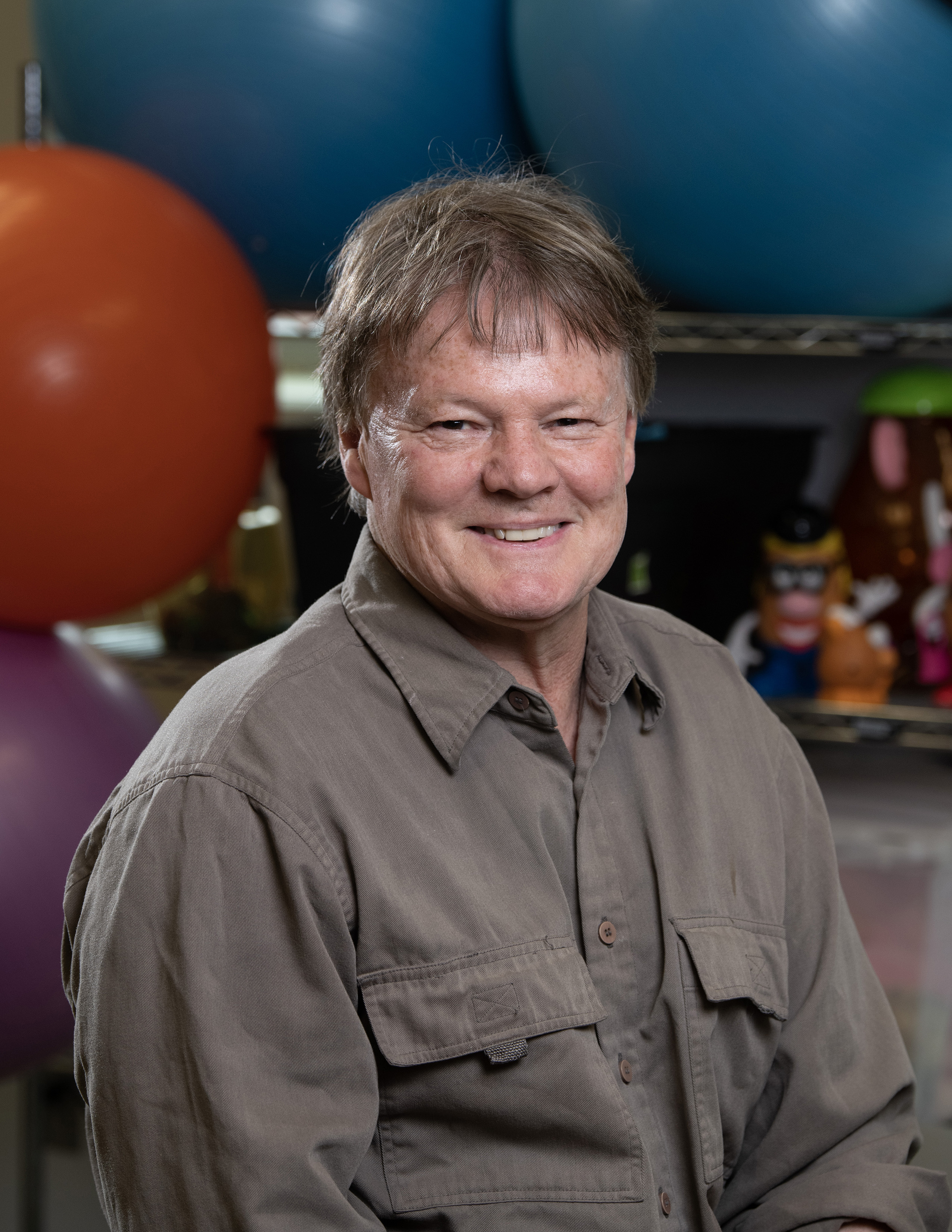 A man smiles at the camera while seated in front of colorful exercise balls.