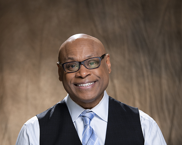 A headshot of a bald man waiting black rimmed glasses, a black collared shirt, a blue plaid tie and a black vest is shown.