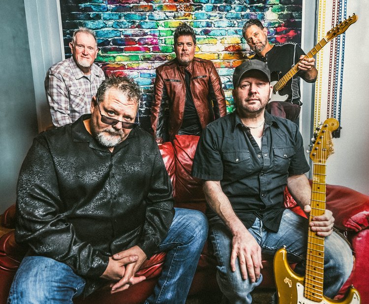 Five men are seated around a red leather couch with a rainbow-colored brick wall behind them. Two of the men hold guitars. 