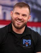 A brown haired man with facial hair wearing a black collared shirt and a smile is shown. He is looking off camera.