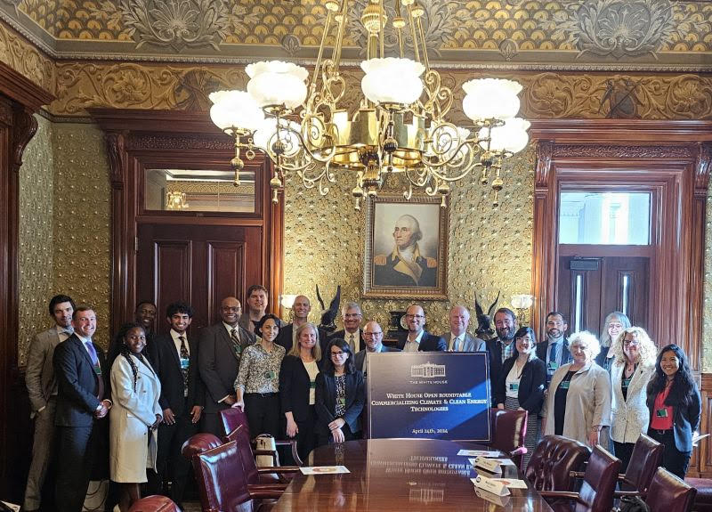 A group of people is shown together standing at the end of a board room table with a sign that indicates it's a roundtable discussion on commercializing climate and clean energy technology.