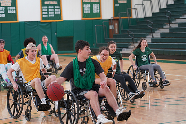 Students in wheelchairs are shown on a basketball court. One dribbles a basketball. 
