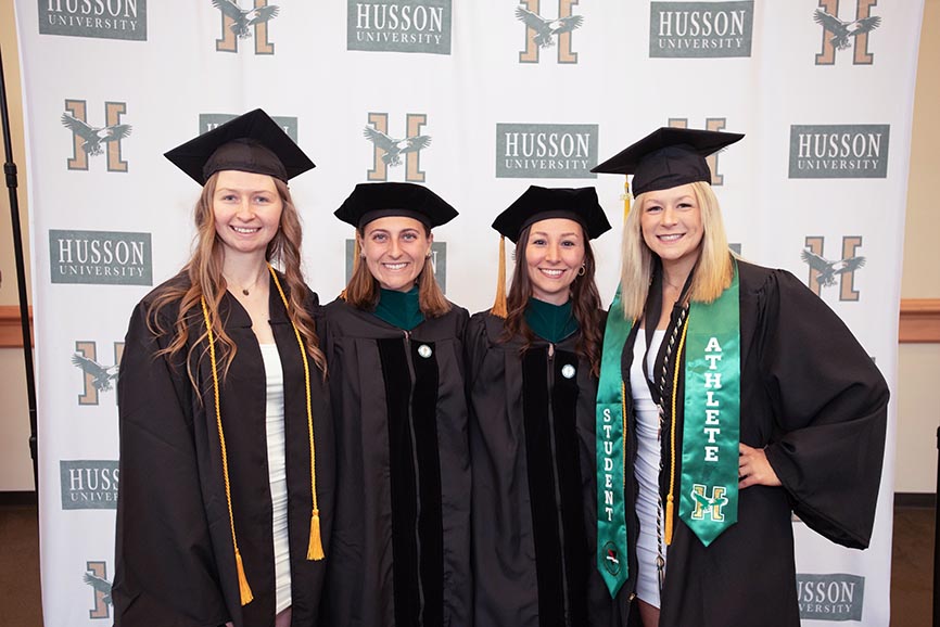 Graduates stand in front of a banner in commencement regalia