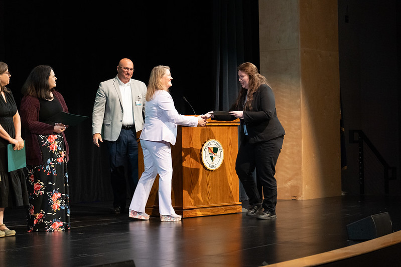 A group of people are shown on a stage. A blond woman is shaking the hand of another woman.