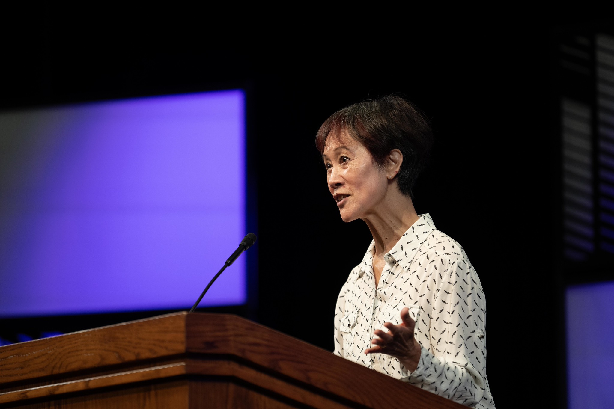 A woman stands at a podium speaking to an audience.
