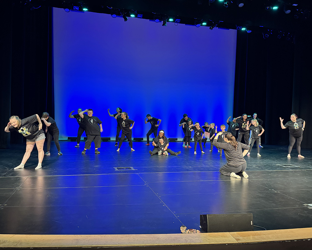 A group of adults and children dance on a stage.