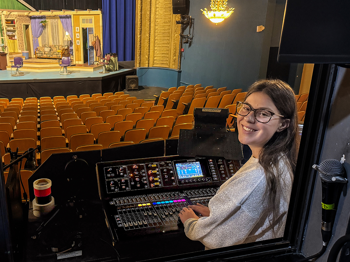 A young woman in glasses looks at the camera. She is seated at a board with sliding controls and a lit stage is in the background.