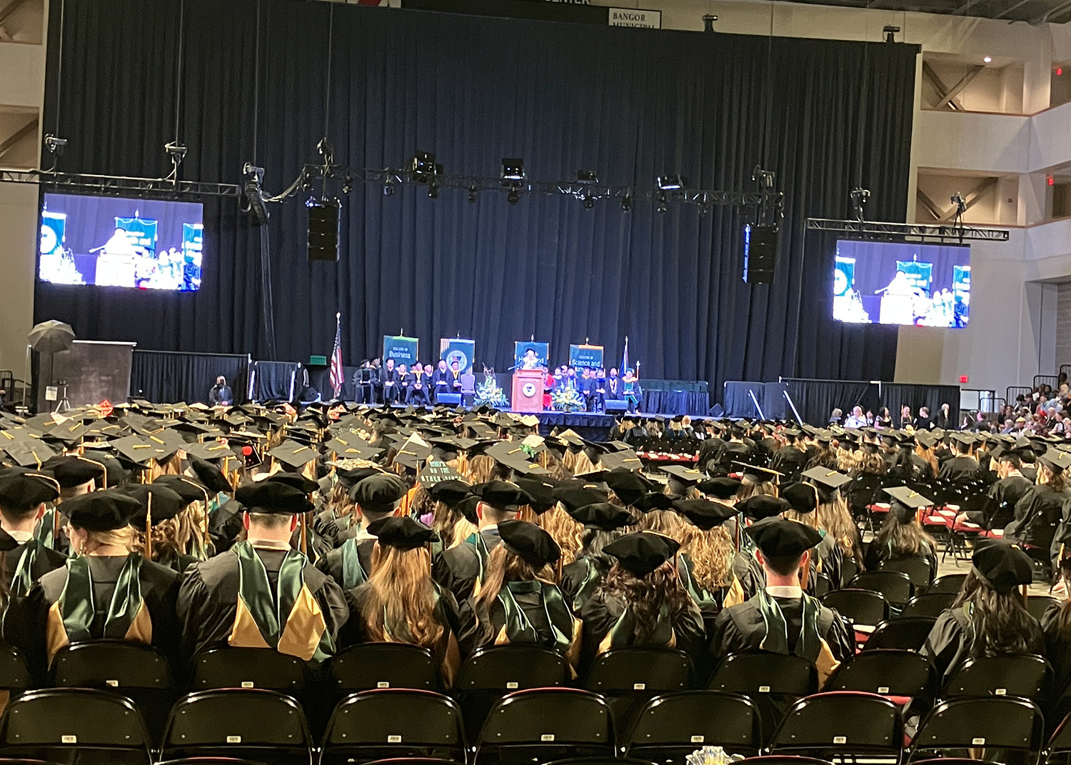 Rows of people in caps and gowns facing a stage with people on it is shown.