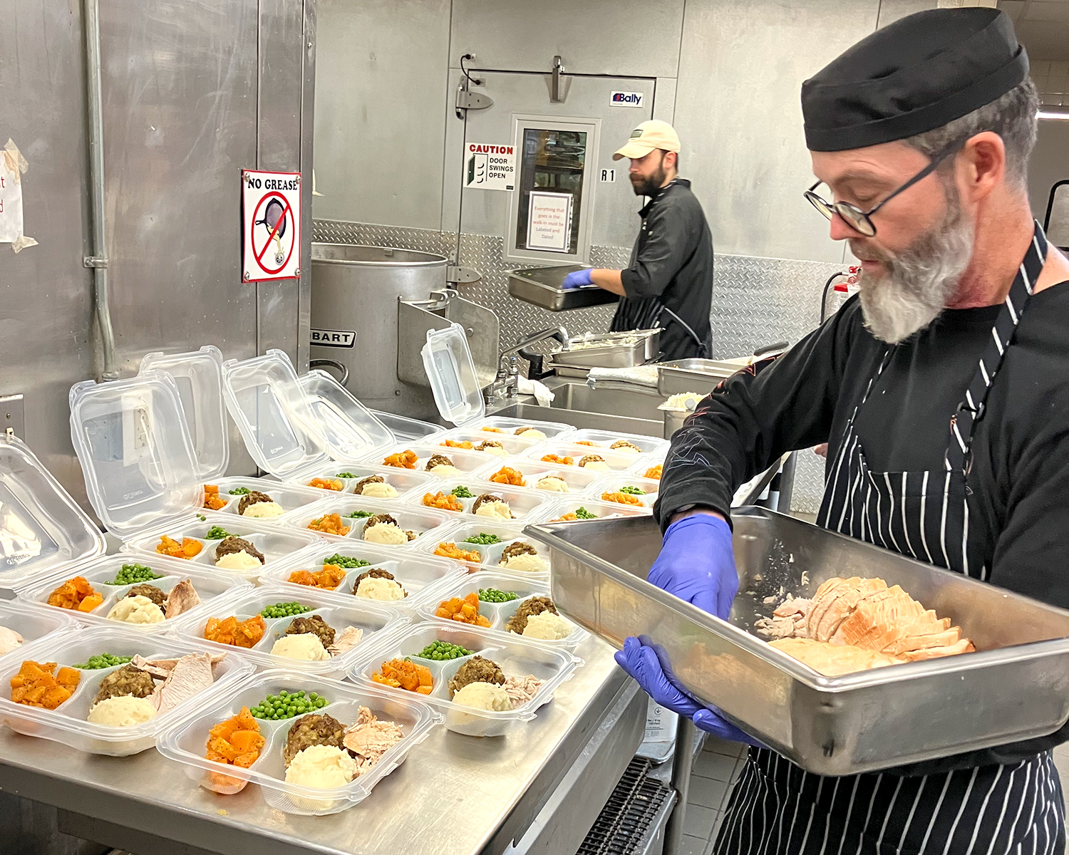 A man in a black chef's uniform adds food to plastic containers containing Thanksgiving meals.