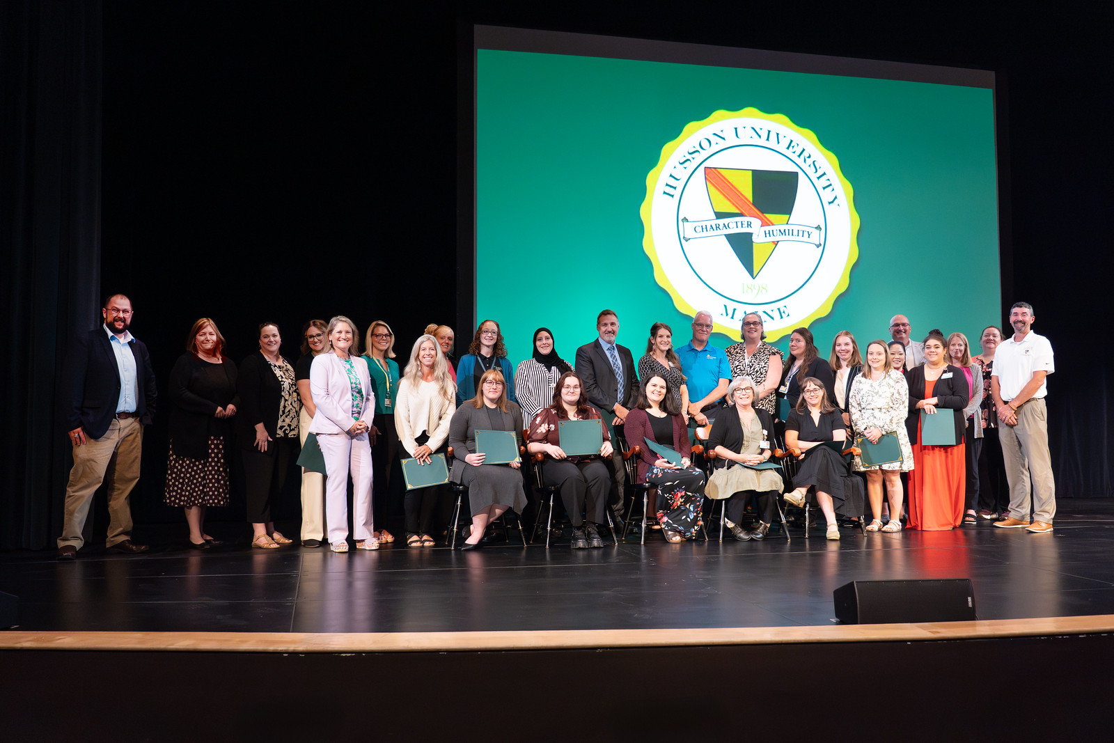 A large group of people holding folders are shown on a stage.