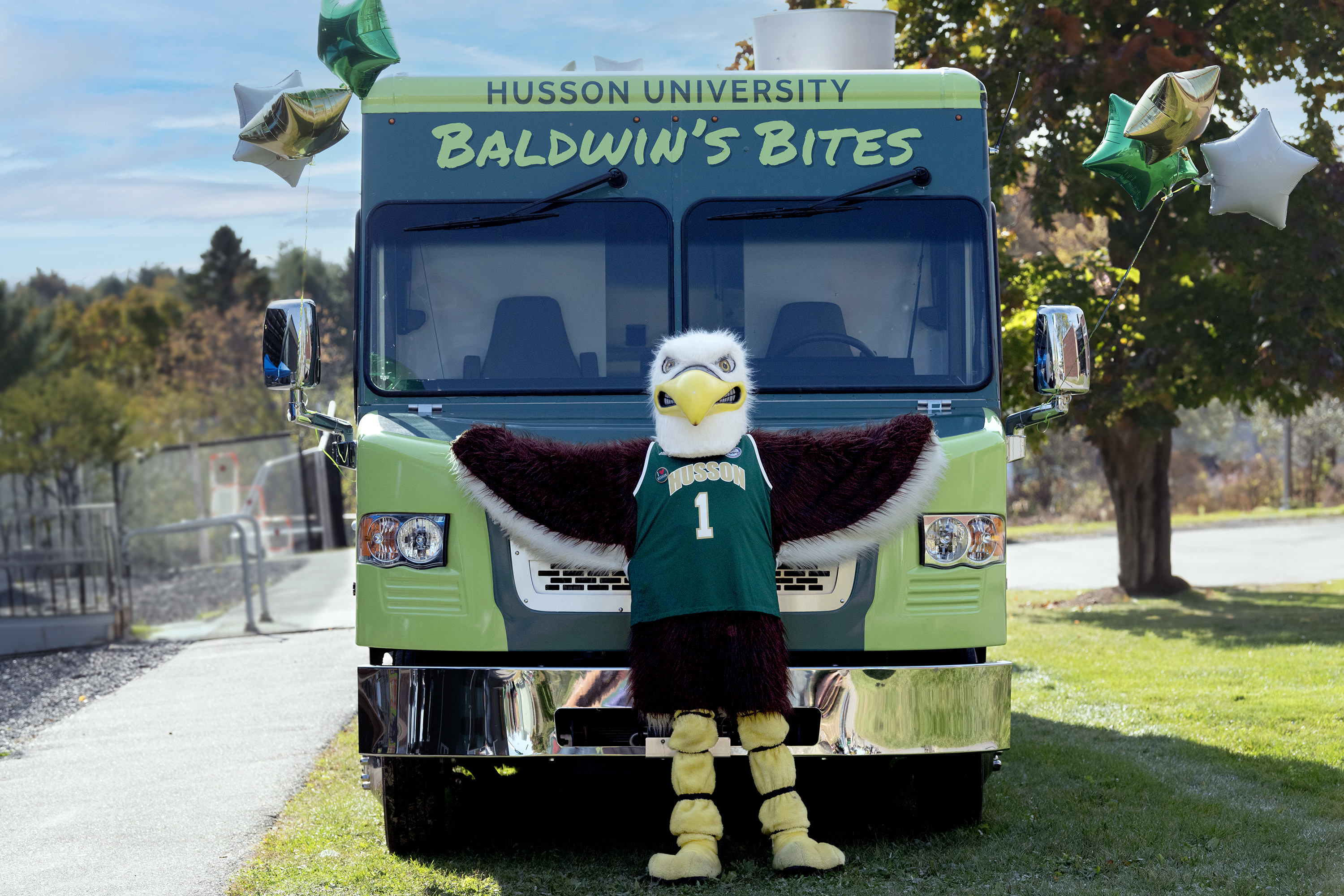 An Eagle mascot wearing a Husson jersey stands with his wings extended in front of a green box truck that says "Baldwin's Bites."