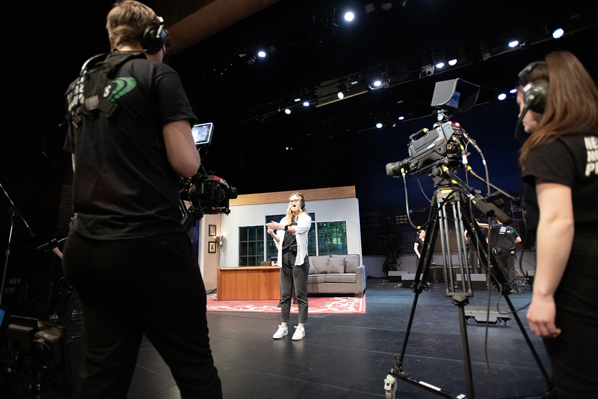 This image shows a stage with a television set and cameras. A young woman wearing a headset, a white collared shirt over a black shirt and black pants gestures to others on the stage.