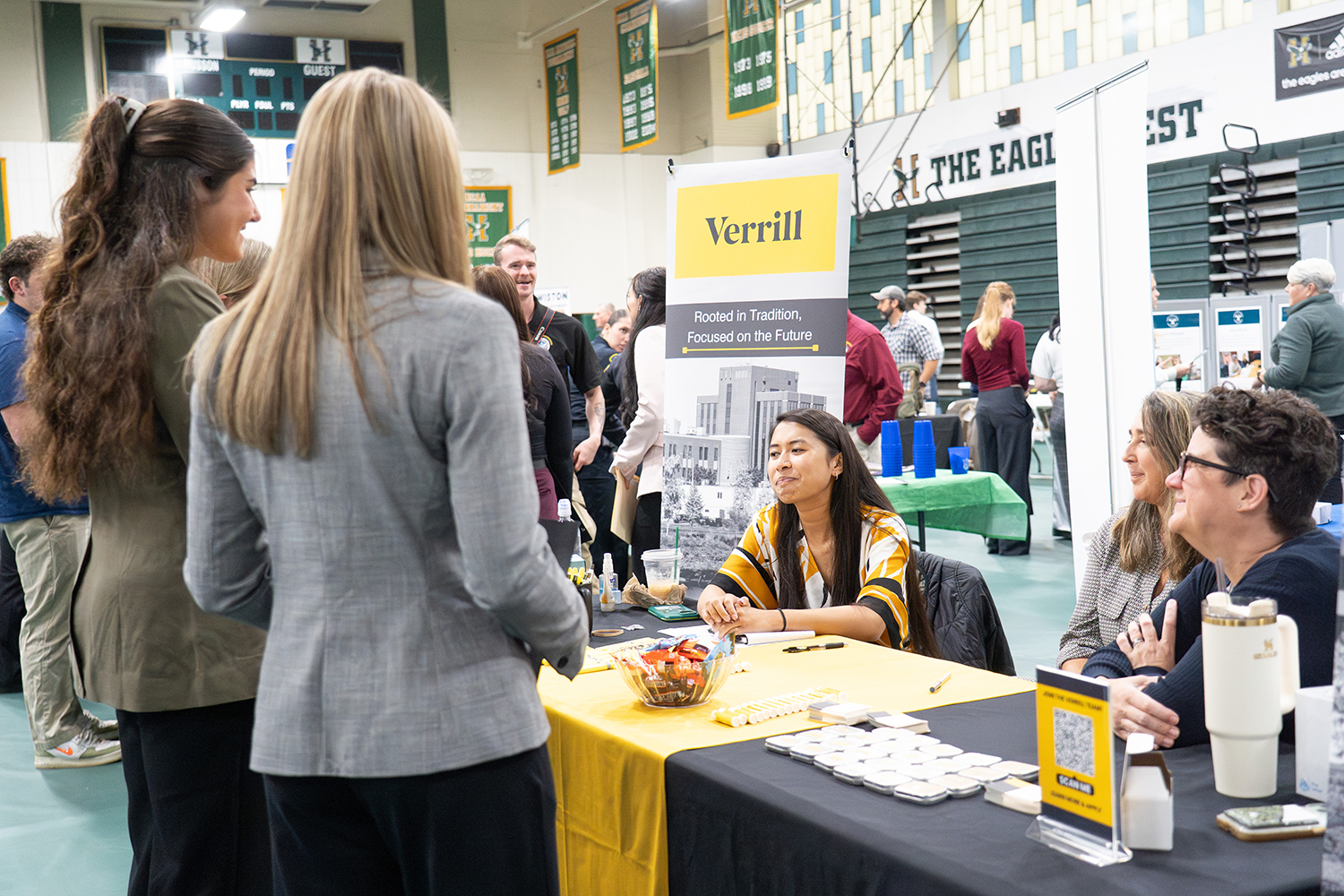 Two young women speak to three seated woman at a table at a career fair.