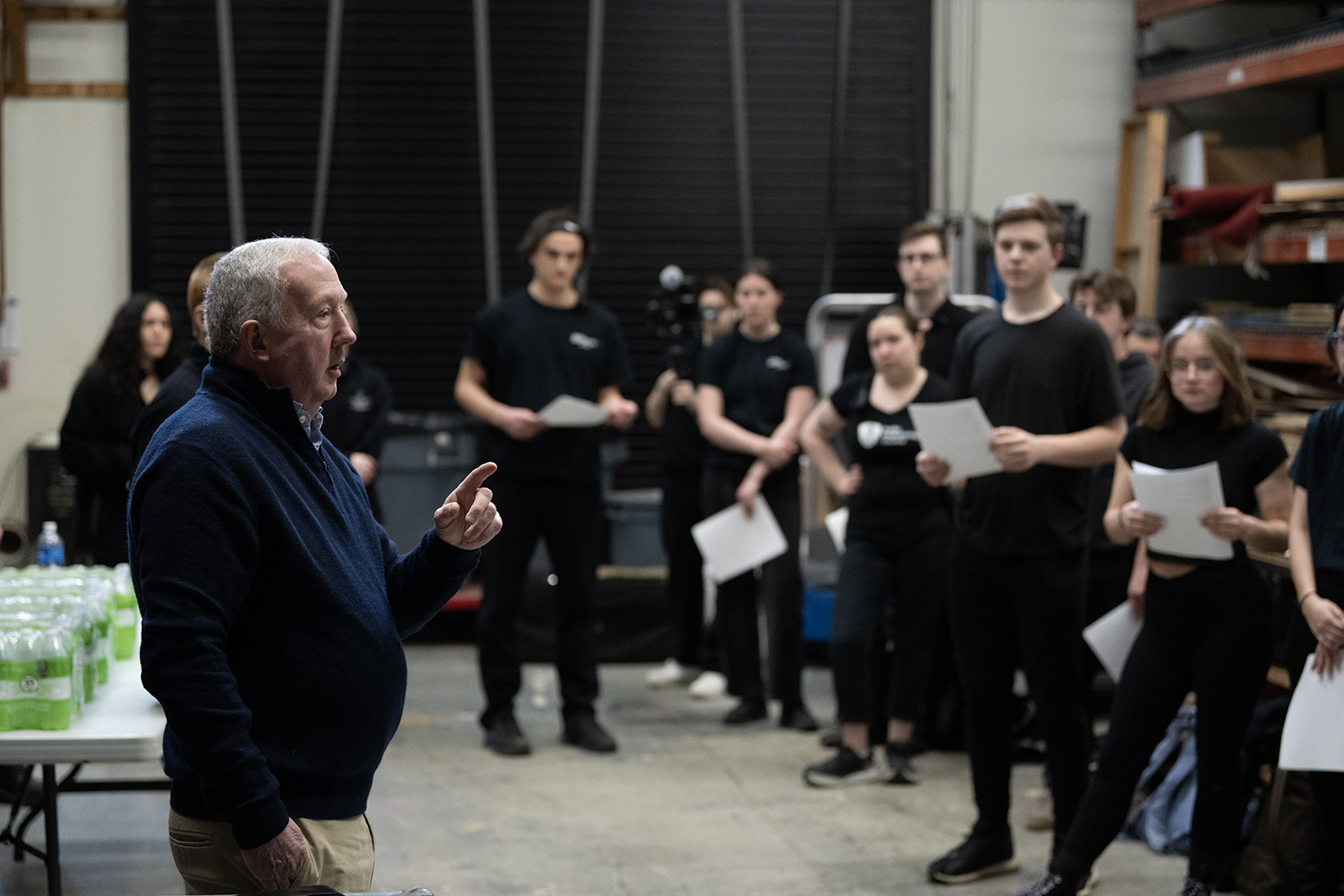 A group of people wearing all black. stand in a semi-circle around an older man who is talking.