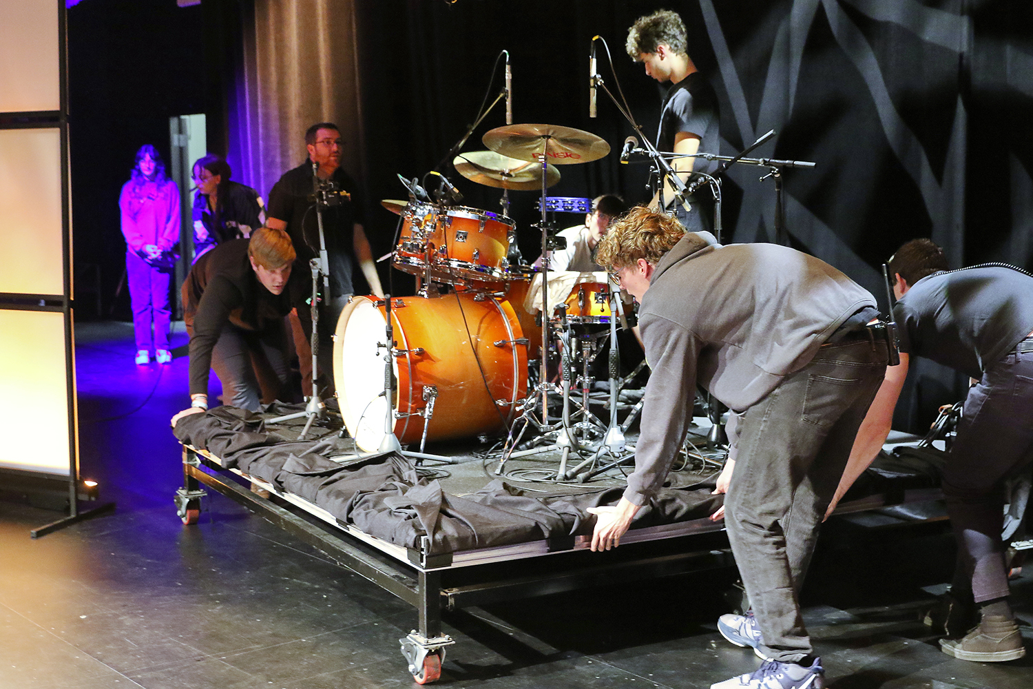 Several people move a set piece with drums on it on a stage.