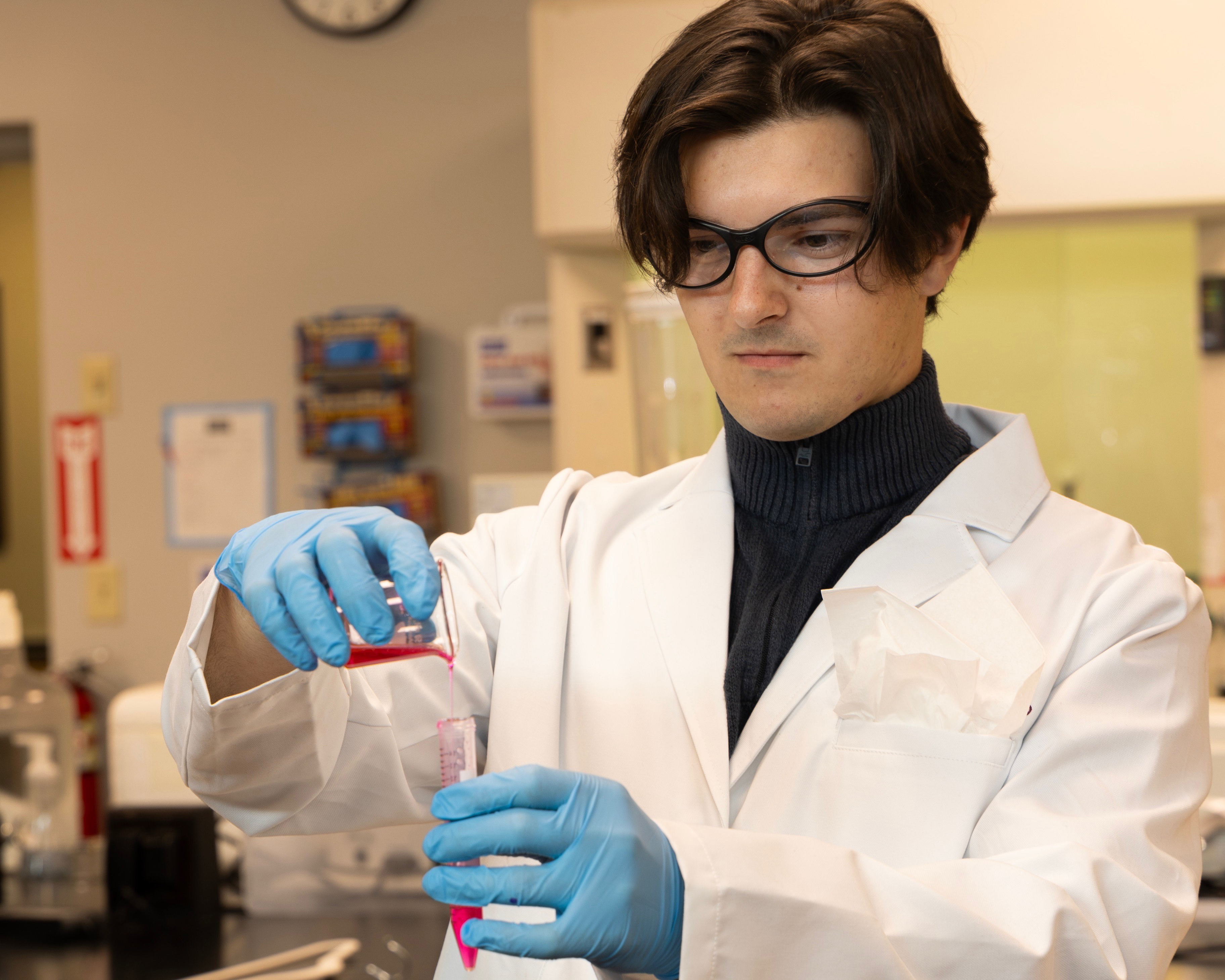 A man in a white lab coat measures liquid from one vessel to another.