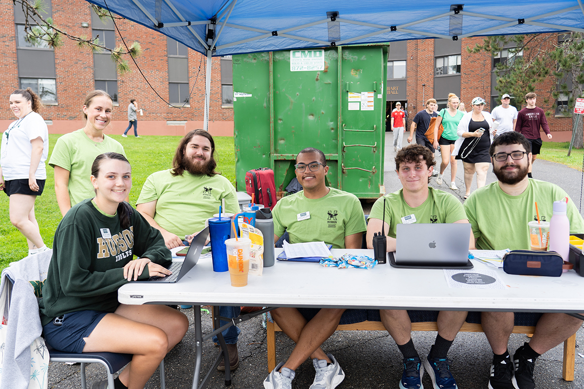 Smiling young adults sit at a table under a tent in this campus photo.