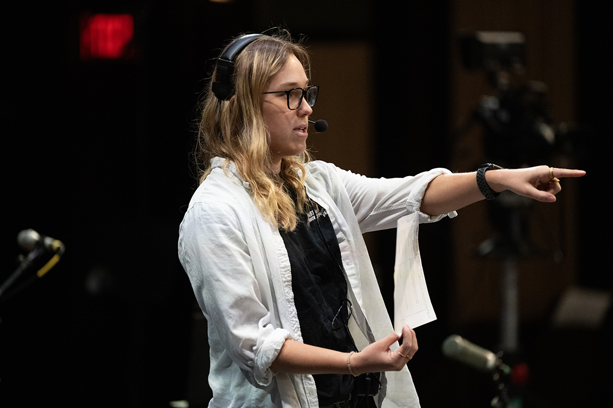 This photo shows a closeup of a young woman with a headset on wearing a white collared shirt over a black shirt.