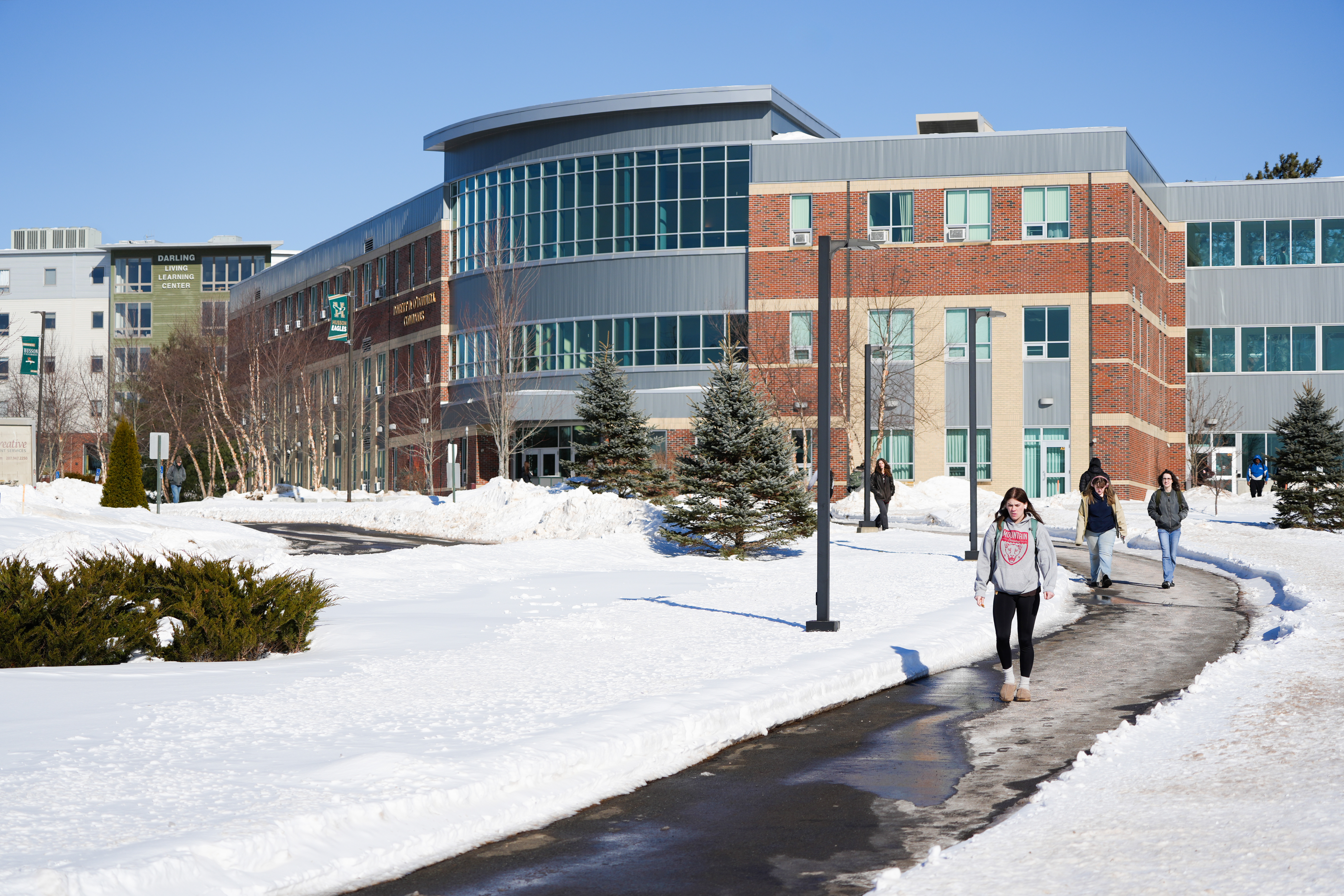 Students walk the path next to Husson University's O'Donnell Commons. There is snow on the ground and the sun is shining bright. 