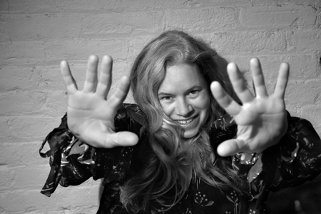 Natalie Merchant poses for a black and white photo. She stands in front of a white brick wall and holds her hands out in front of her while wearing a black floral shirt. 