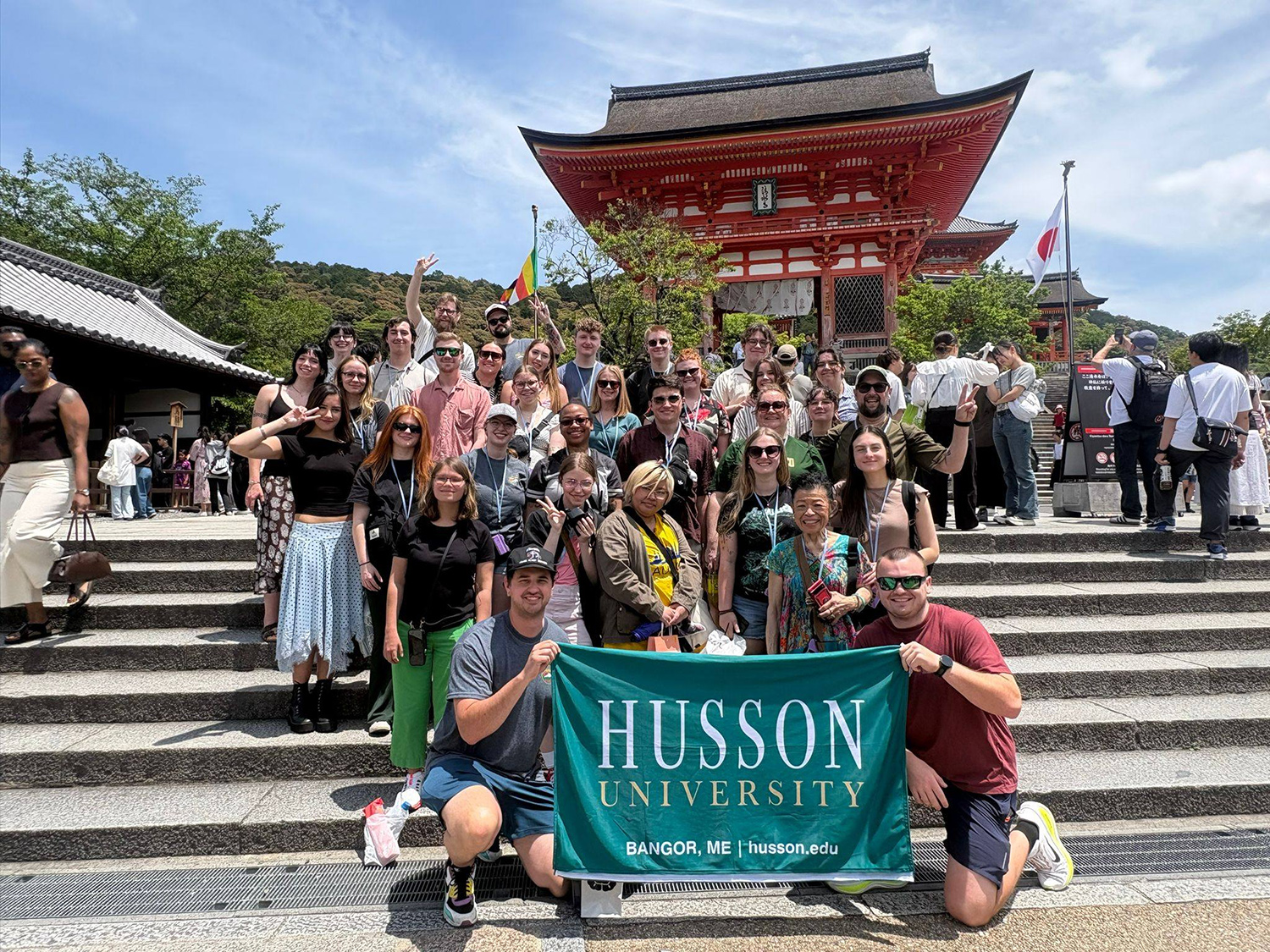 A large group of people holding a "Husson University" banner stand in front of a Japanese building.