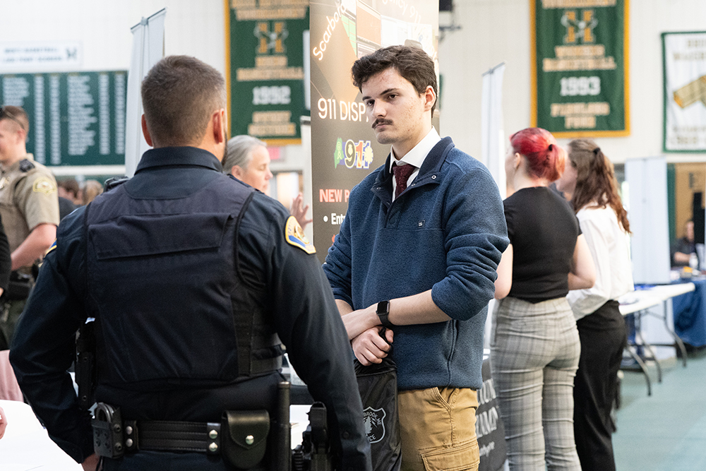 A young man with a mustache speaks to a member of law enforcement at a career fair. Other people and tables are nearby.