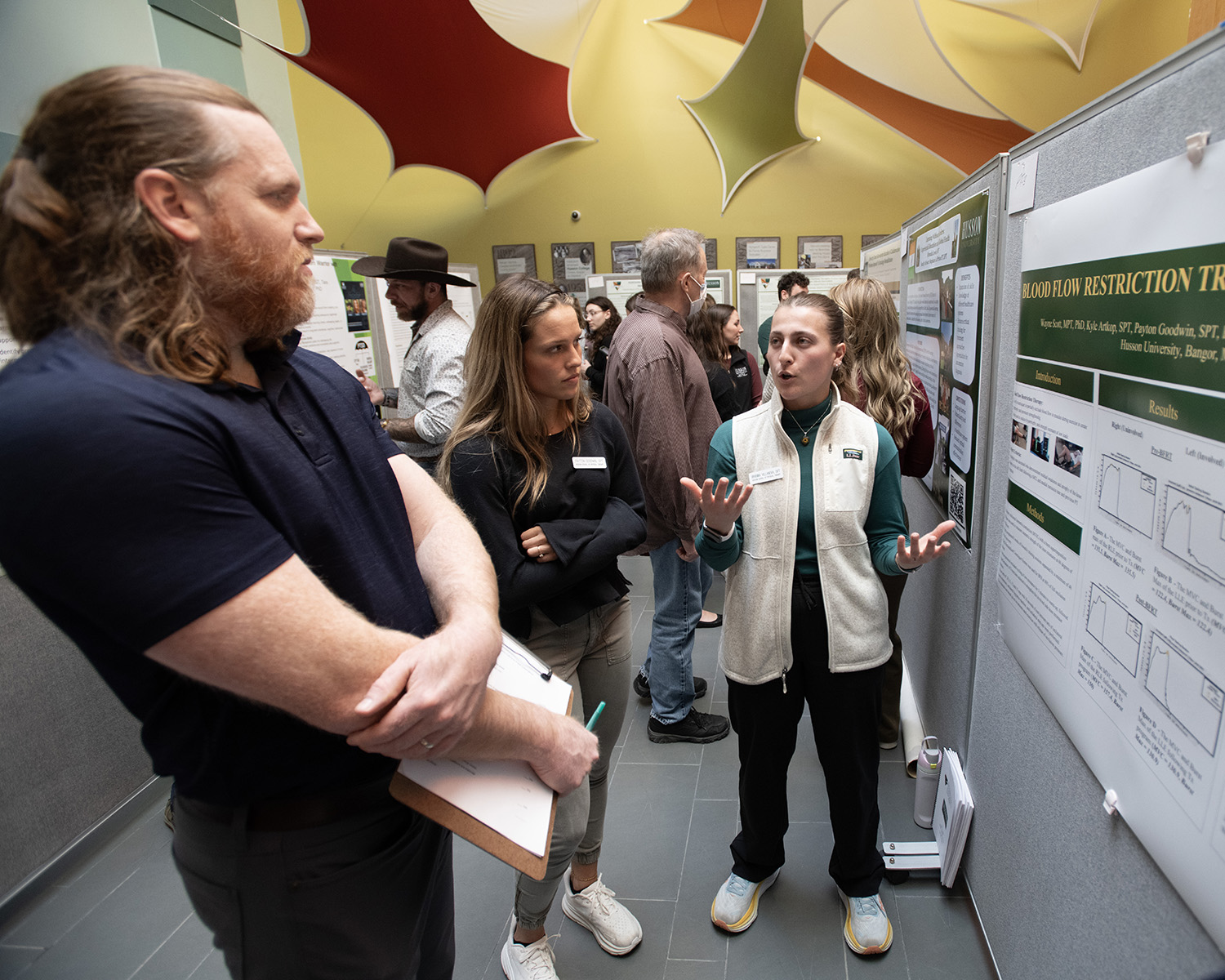 A young woman speaks about her research to a man and woman. They are standing next to a research poster.