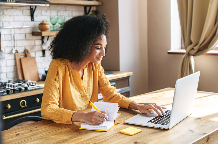 woman working on a computer and writing in a notebook while sitting at a table in a kitchen