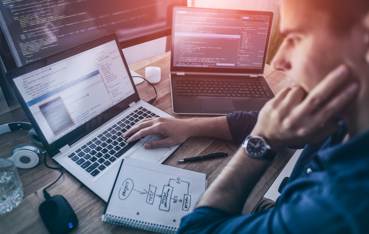 Man working on multiple computers while also looking at a graph drawn on a notebook.