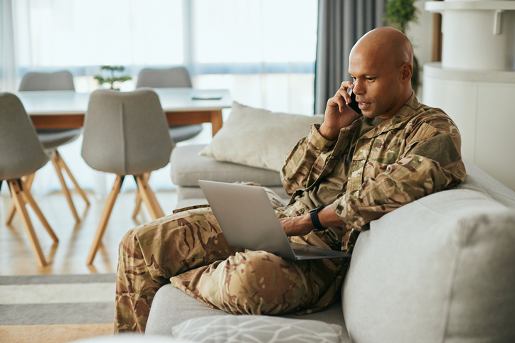 Military veteran sitting using a laptop computer and cell phone while sitting on a couch.