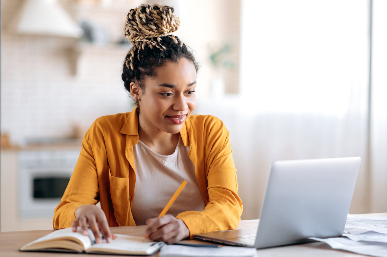 A female student taking notes in a notebook while looking at her laptop during an online class.