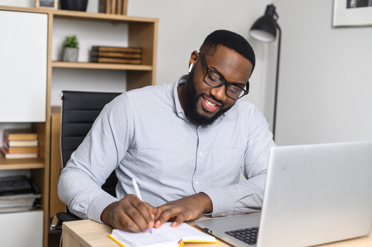 Online business student writing notes while taking an online class on a laptop at a desk.